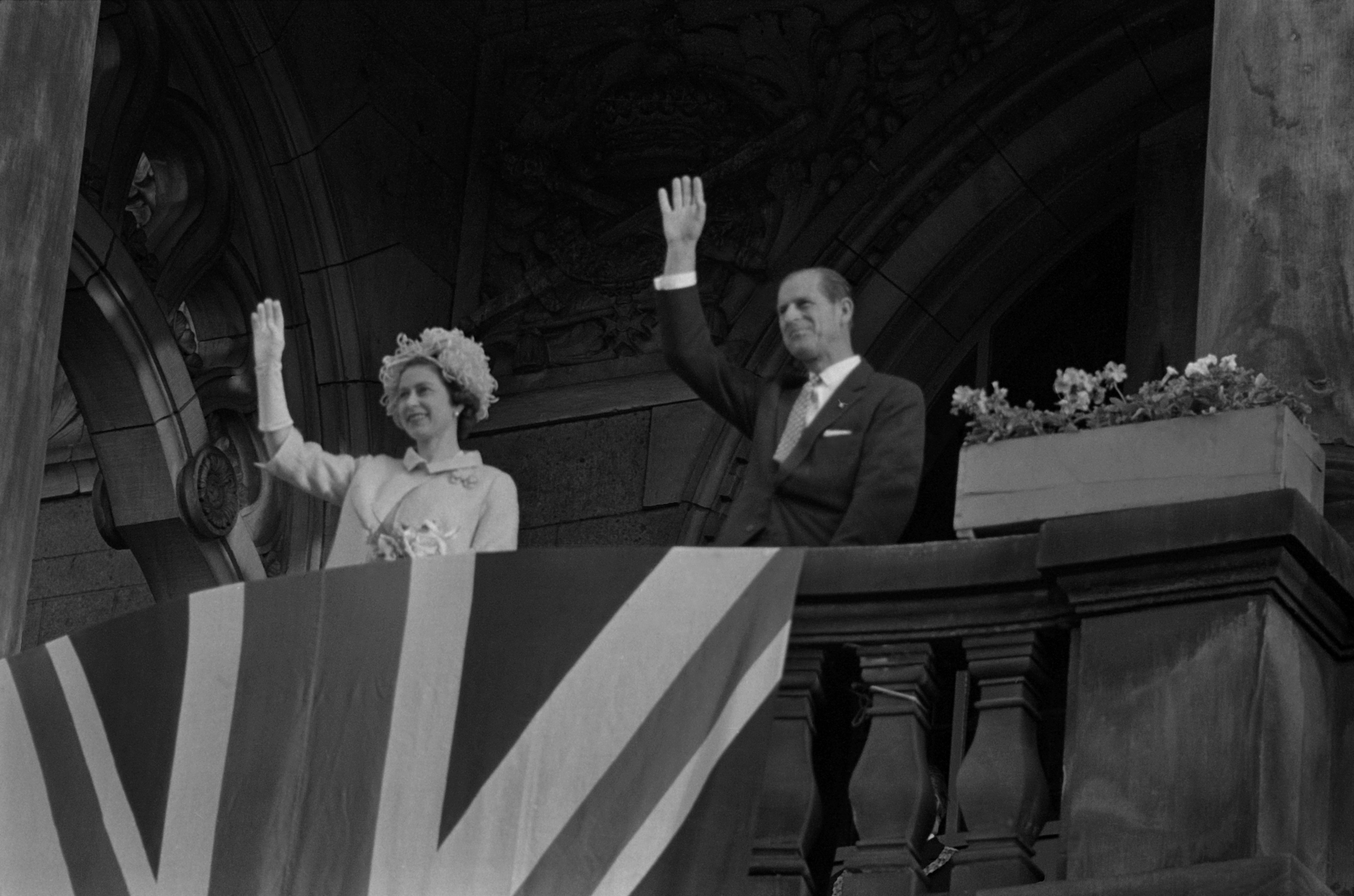 Queen Elizabeth II  and Prince Philip, Duke of Edinburgh, wave to the crowds from a balcony behind a draped Union Jack flag on May 28, 1965, in West Berlin, West Germany.